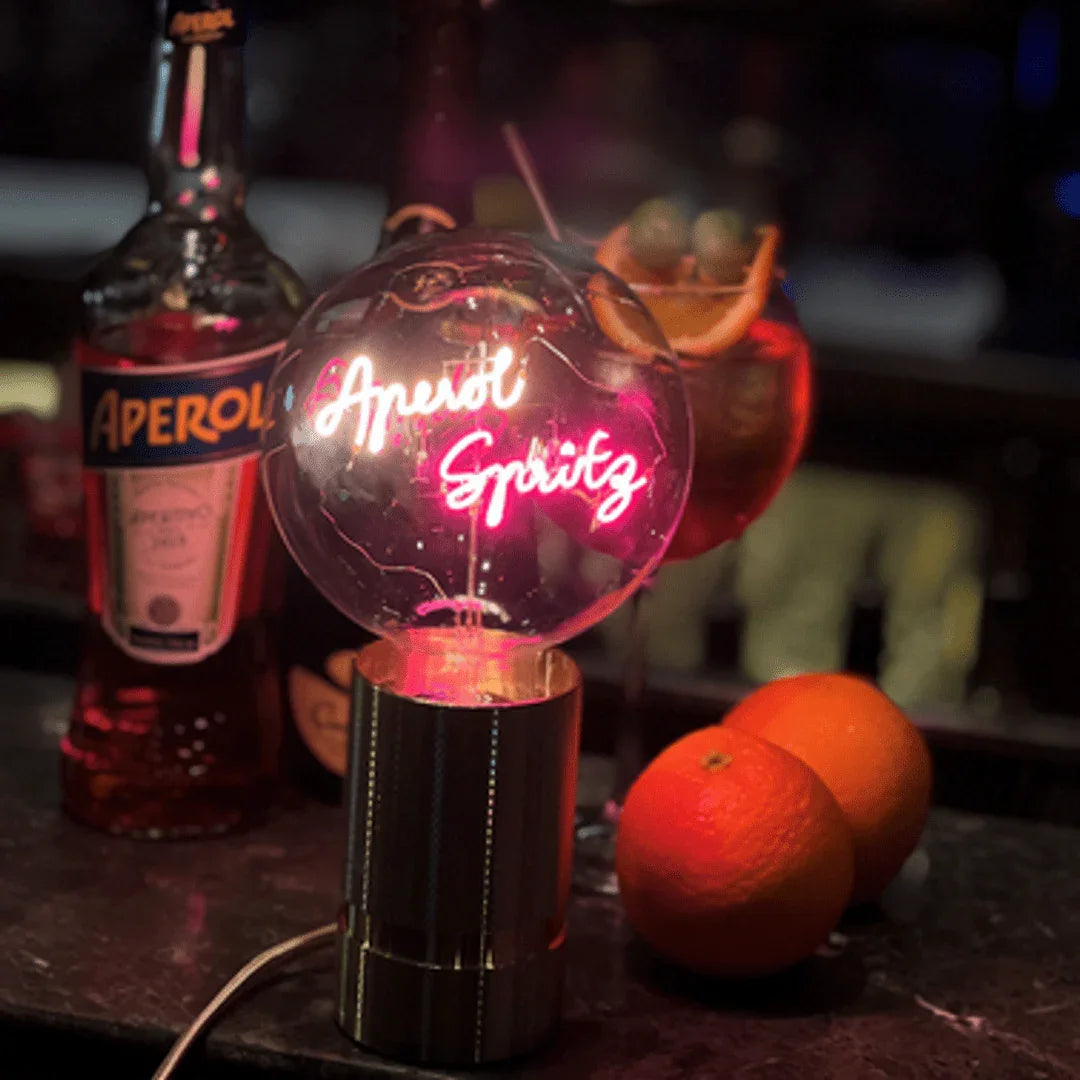 Ampoule LED design avec message lumineux Aperol Spritz, posée sur un bar, oranges et bouteille en fond