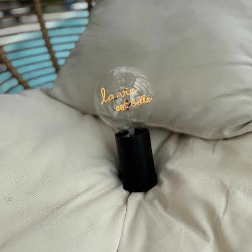 Ampoule LED à message la vie est belle sur socle noir, posée sur un coussin beige